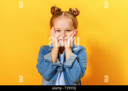 Closeup ritratto di un simpatico attraente piccola ragazza su sfondo giallo. Bambino mani viso e guarda nella telecamera. Foto Stock