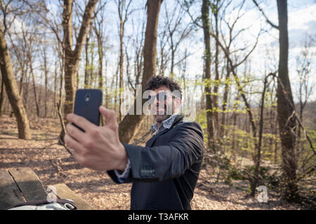 Un giovane uomo, utilizza lo smartphone per effettuare un selfie, all'aperto in natura. con la sua lingua, facendo una faccia buffa. braccio allungato nella parte anteriore. Foto Stock