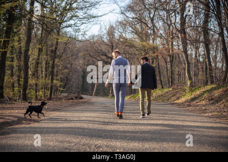Vista posteriore, due gay uomo a camminare nella foresta, su strada asfaltata. Insieme tenendo le mani. Chiamare per il loro cucciolo di cane. Foto Stock