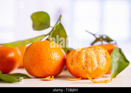 Il gruppo di cinque intere fresche arancione mandarine con foglie verdi un frutto è metà sbucciata con finestra blu in una cucina bianca Foto Stock