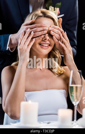 Messa a fuoco selettiva di felice sposa con gli occhi chiusi da stallieri mani isolato su nero Foto Stock