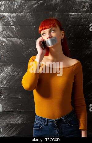 Quadro concettuale. I giovani i Capelli rossi ragazza cerca di parlare al telefono con sigillata con nastro adesivo con le labbra in modo che essa mantiene il silenzio Foto Stock