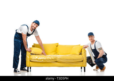Due movers guardando la fotocamera durante l'avvolgimento divano giallo con una bobina di film estensibile su bianco Foto Stock