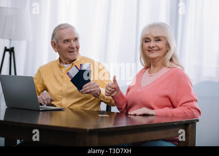 Senior uomo trattenere aria biglietti e passaporti vicino a donna che mostra il pollice in alto segno a casa Foto Stock