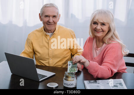 Coppia senior seduta a tavola con laptop e guardando la telecamera mentre donna mettere soldi in un barattolo di vetro con il termine "pensione" scritte Foto Stock