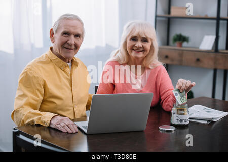 Coppia senior seduta a tavola con laptop e guardando la telecamera mentre donna mettere soldi in un barattolo di vetro con il termine "pensione" scritte Foto Stock