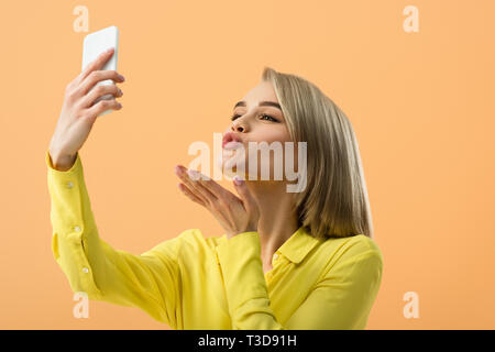 Bella ragazza bionda di inviare aria kiss tenendo selfie isolati su Orange Foto Stock