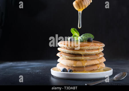 Dolci fatti in casa pila di pancake con colata di miele, ai mirtilli freschi e menta in una moderna piastra su sfondo nero. Spazio per il testo. Foto Stock