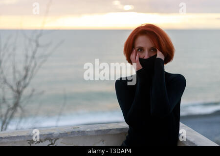 Ritratto di una giovane donna con i capelli rossi con un inquieto guardare sullo sfondo del mare e del tramonto in serata primaverile Foto Stock