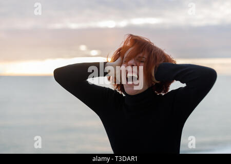 Giovane donna con i capelli rossi che stringe la testa e urlando a gran voce da heartache sullo sfondo del mare e del tramonto Foto Stock