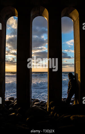 Tre aperture attraverso le grandi colonne di cemento di un ponte mostra la scena di un oceano al tramonto. Un uomo si appoggia contro la colonna di destra guardando a bea Foto Stock