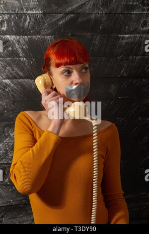 Quadro concettuale. I giovani i Capelli rossi ragazza cerca di parlare al telefono con sigillata con nastro adesivo con le labbra in modo che essa mantiene il silenzio Foto Stock