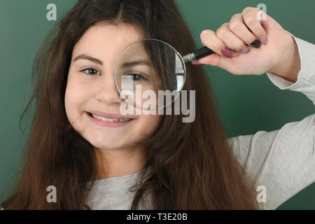 Sorridente Little Schoolgirl su sfondo verde . Giovane ragazza giocoso detiene una lente di ingrandimento in mano Foto Stock