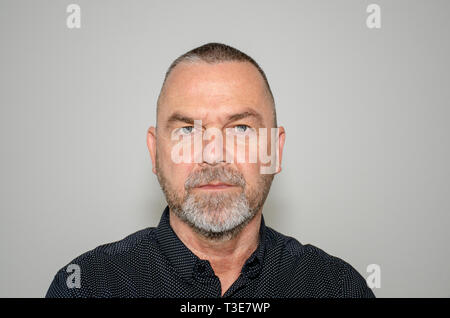 Intensa determinata barbuto uomo di mezza età fissando la telecamera con una risoluta grim espressione su un grigio di sfondo per studio Foto Stock