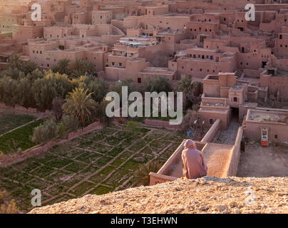 Un uomo seduto su di una collina che guarda verso il Ksar Aït-Ben-Haddou, Aït Benhaddou‌, provincia di Ouarzazate, Drâa-Tafilalet, Marocco, Africa. Foto Stock