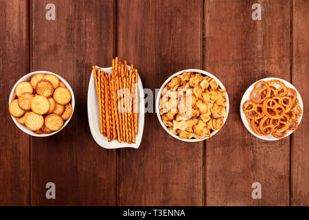 Il salato crackers, bastoncini, salatini e goldfishes, girato dalla parte superiore su di un legno scuro dello sfondo con copia spazio. Party Mix snack Foto Stock