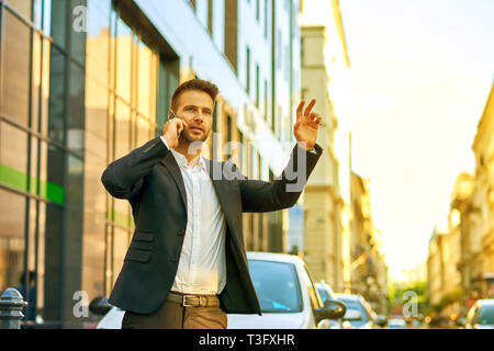 Un bel giovane imprenditore in piedi di fronte ad un edificio di uffici, parlando sul suo telefono cellulare e agitando per un taxi con la mano Foto Stock