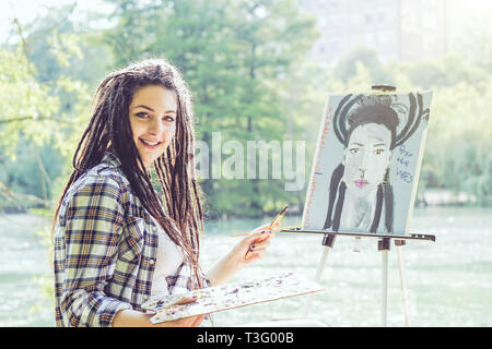 Artista giovane ragazza di verniciatura di un ritratto di auto in un parco vicino al lago - Pittore donna con dreadlocks acconciatura lavorando sulla sua arte in città per esterno Foto Stock