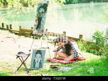 Artista giovane ragazza bozza di disegno di prossima pittura su parco vicino al lago - Pittore donna con dreadlocks acconciatura lavorando sulla sua arte in città per esterno Foto Stock