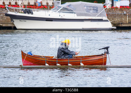 Uno steward che indossa una giacca a vento e giallo sou'wester hat si siede in un ceppo di legno barca a remi ormeggiate nel fiume Tamigi in un giorno di pioggia a Henley Royal Regatta Foto Stock
