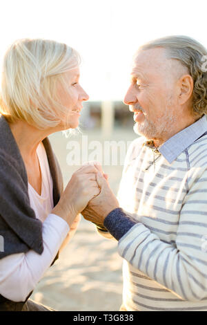 Anziani bionda moglie caucasica holding hands senior marito e seduto sulla spiaggia di sabbia. Concetto di felice pensionati su pic-nic. Foto Stock