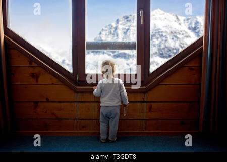 Bambino bambino in piedi di fronte a un grande porte francesi, appoggiandosi contro di essa guardando al di fuori di una natura innevata, giocando con i giocattoli Foto Stock