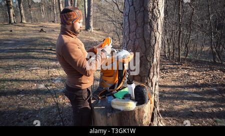 Escursioni a tema e viaggi. Un turista caucasica uomo estrae un arancio zaino, prende le sue cose e li mette su un moncone nella foresta. Un equipaggiamento Foto Stock