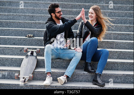 Cool multirazziale giovane seduto sulle scale con longboard e dare alta cinque di ogni altro. Foto Stock