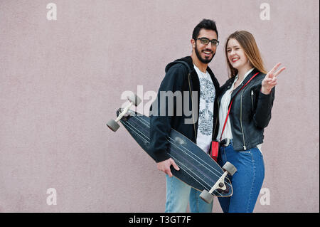 Cool multirazziale matura in posa contro la parete rosa con longboard. Foto Stock