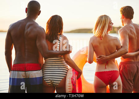 Specchietto di due giovani coppie in costume da bagno in piedi a braccetto insieme a guardare il tramonto su un lago Foto Stock