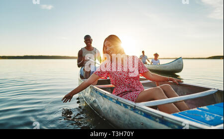 Sorridente giovane donna immergendo la mano in acqua di lago mentre paddling una canoa con il suo fidanzato e amici su un tardo pomeriggio estivo Foto Stock