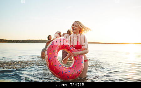 Ridendo giovane donna in un bikini in possesso di un anello di nuotare con gli amici gli schizzi in background mentre si divertono insieme a un lago Foto Stock