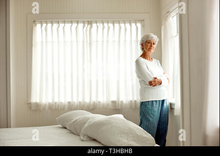 Infelice donna matura in pigiama sta con le braccia incrociate davanti a una camera da letto finestra come lei pone per un ritratto. Foto Stock