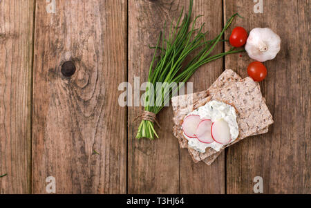 Fette biscottate con quark e di erba cipollina fresca, ravanelli e pomodori su una tavola in legno rustico - una sana colazione con erbe fresche Foto Stock