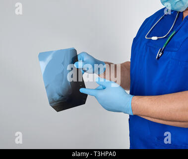 Dottore in uniforme blu e sterili Guanti in lattice trattiene e esamina X-ray di osso della gamba e sfondo grigio Foto Stock