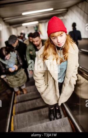 Ragazza adolescente con Red Beanie e cuffie in ear su scala mobile a una stazione della metropolitana, Colonia, nella Renania settentrionale-Vestfalia, Germania Foto Stock