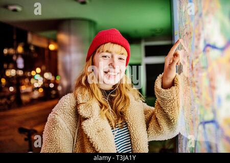 Ragazza adolescente, con un red beanie e cuffie nel suo orecchio in piedi di fronte ad una mappa della città di notte, Colonia, nella Renania settentrionale-Vestfalia, Germania Foto Stock