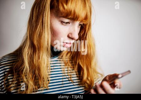 Ragazza adolescente, i capelli rossi, appare spaventato al suo smartphone, studio shot, Germania Foto Stock