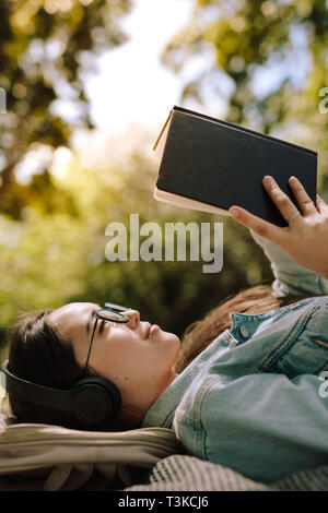 In prossimità di una giovane donna che giace a terra e la lettura di un libro di indossare le cuffie. La donna in un'atmosfera rilassata, la lettura di un libro che giace all'aperto durante l'ascolto Foto Stock