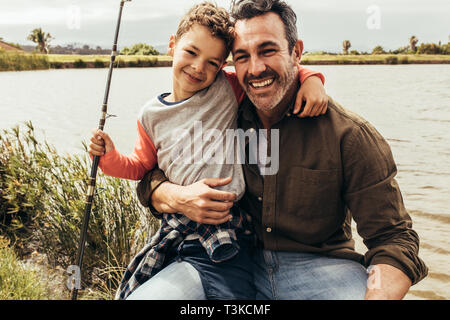 Sorridente padre e figlio seduto accanto a un lago in possesso di una canna da pesca. Chiudere da padre e figlio in un giorno fuori di trascorrere del tempo insieme la pesca in prossimità di un lago Foto Stock