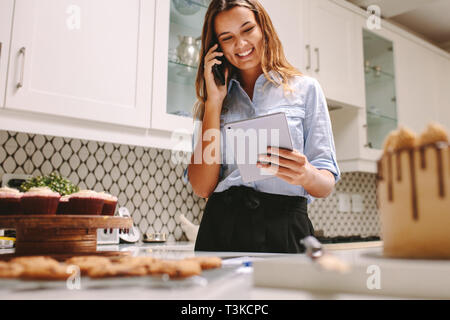 Giovani chef pasticcere pronto per iniziare la sua propria impresa di cottura di torte per l'ordine. Felice la cucina dello chef in cucina sorridente e utilizzando una tavoletta digitale e talkin Foto Stock