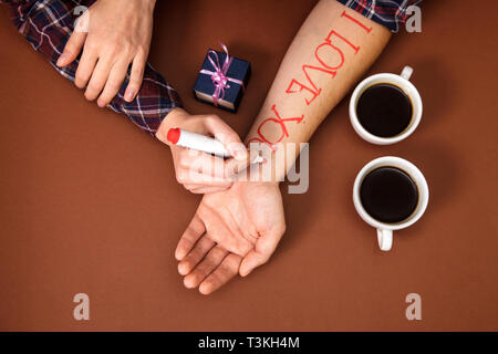 Penna per scrivere una lettera cari. una femmina scrive a mano ti amo su una mano d'uomo. Su uno sfondo marrone ci sono due tazze di caffè e la mano dell'uomo è Foto Stock