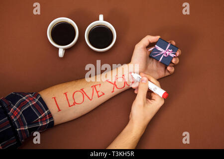 Penna per scrivere una lettera cari. una femmina scrive a mano ti amo su una mano d'uomo. Su uno sfondo marrone ci sono due tazze di caffè e la mano dell'uomo è Foto Stock