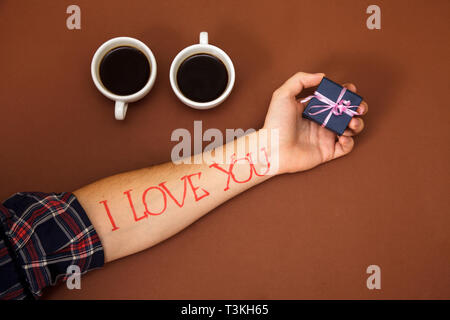 Una mano d'uomo su uno sfondo marrone con lettere ti amo. Un uomo nel palmo della sua mano trattiene una casella blu. Foto Stock