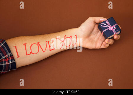 Una mano d'uomo su uno sfondo marrone con lettere ti amo. Un uomo nel palmo della sua mano trattiene una casella blu. Foto Stock