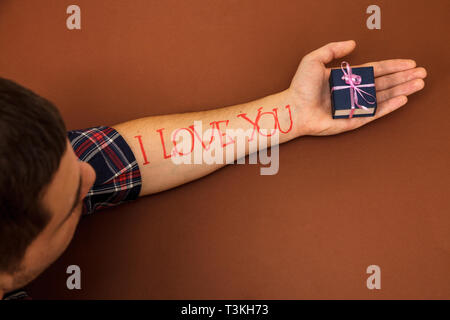 Una mano d'uomo su uno sfondo marrone con lettere ti amo. Un uomo nel palmo della sua mano trattiene una casella blu. Foto Stock