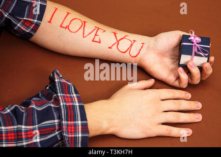 Una mano d'uomo su uno sfondo marrone con lettere ti amo. Un uomo nel palmo della sua mano trattiene una casella blu. Foto Stock