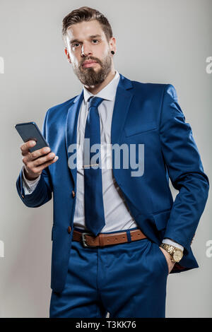 Ritratto di un imprenditore in tuta blu shirt tie golden orologi di lusso smartphone holding guardando la telecamera isolata contro uno sfondo grigio fortuna Foto Stock