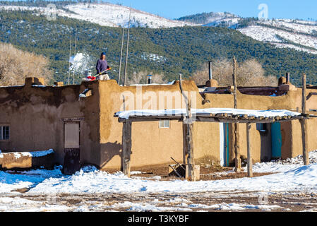 TAOS PUEBLO, Nuovo Messico / STATI UNITI - 19 novembre 2015: l'uomo non identificato pale neve dal tetto di un abitato tradizionale casa di adobe Foto Stock