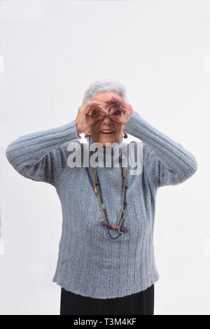 Senior donna guardando attraverso le dita come se si indossano occhiali su sfondo bianco Foto Stock